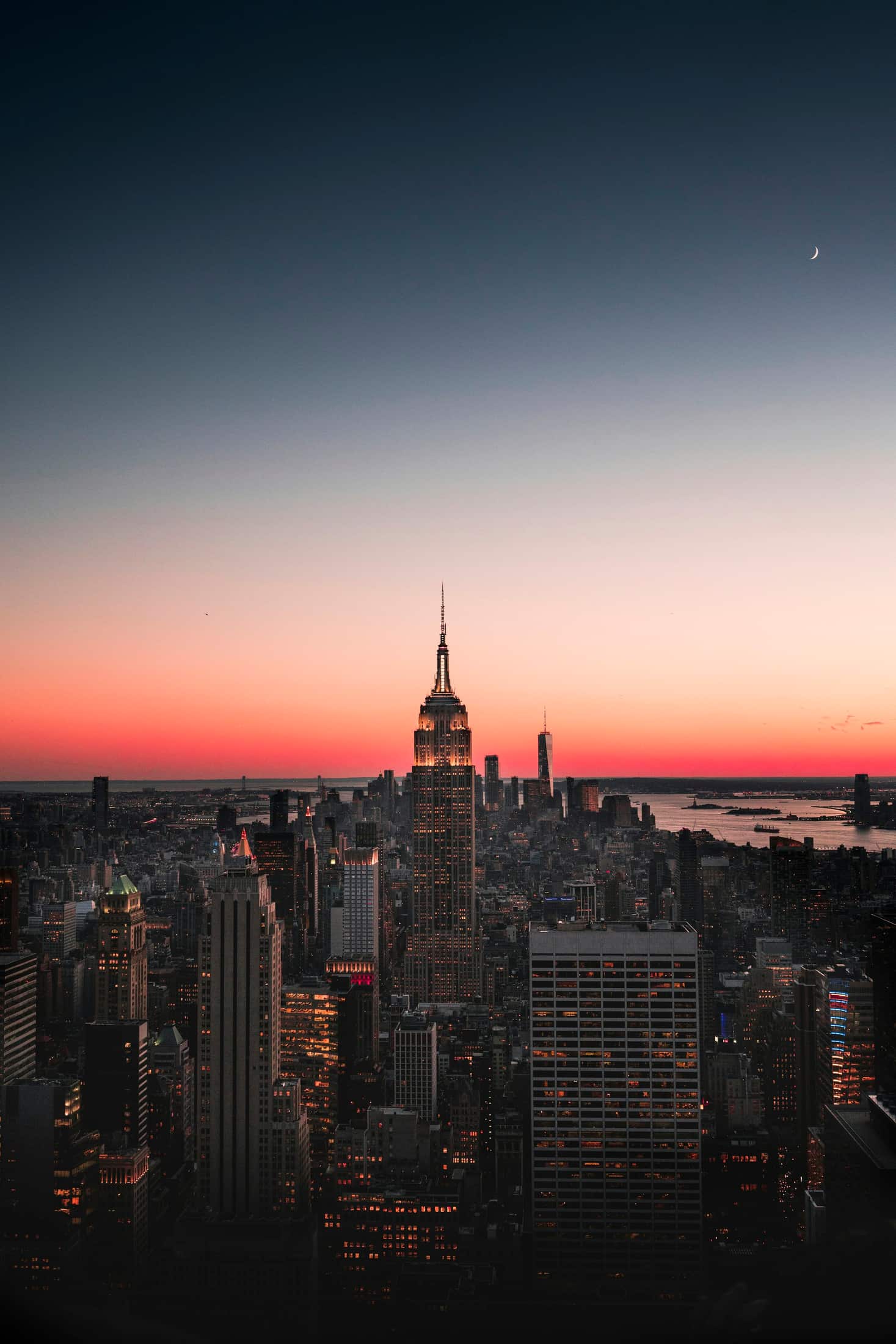 A downtown skyline under a deep dusk sky, a band of ember light along the horizon and a thin crescent moon above